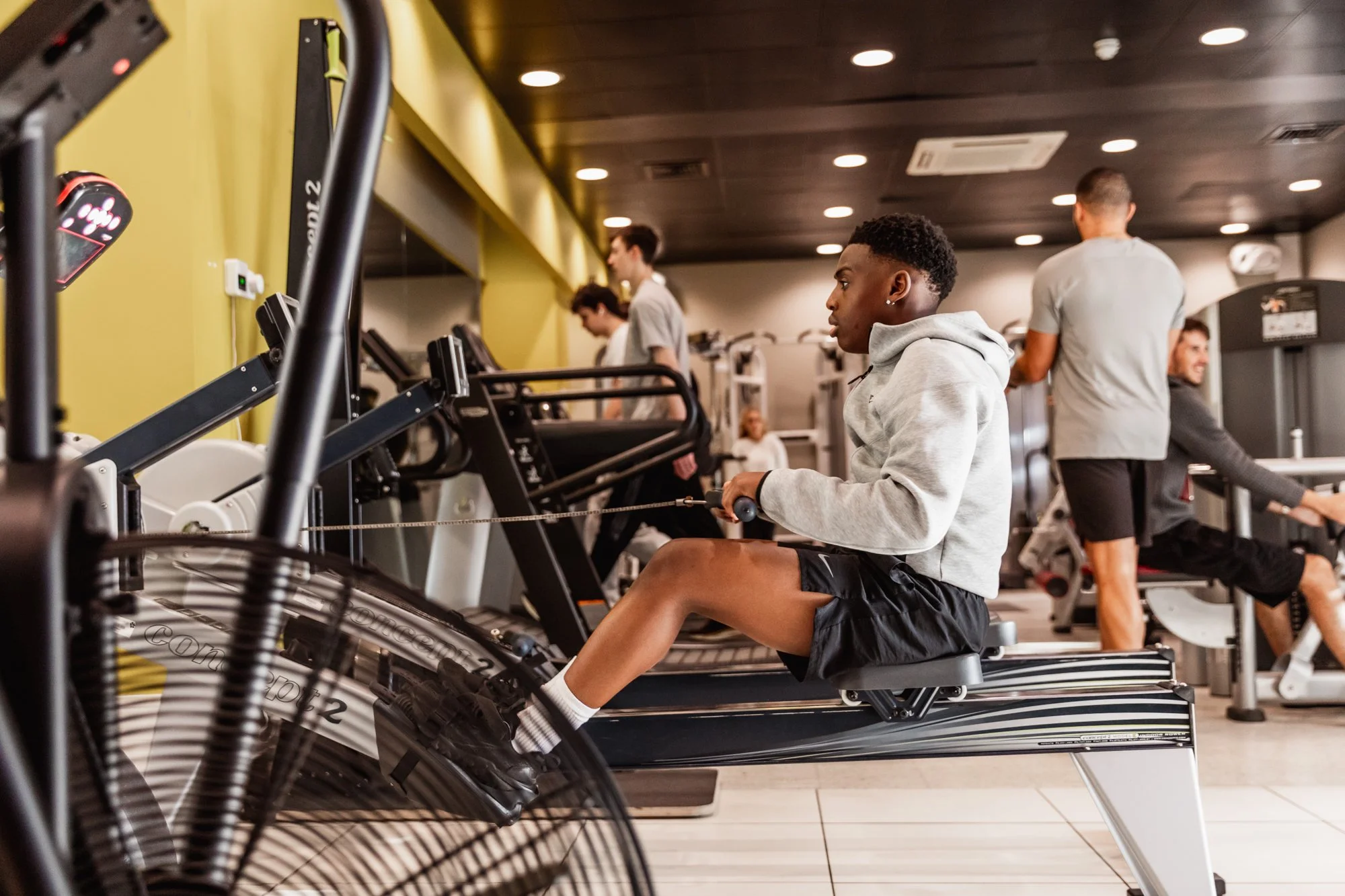 Young man using a rowing machine at the gym with other people working out in the background.