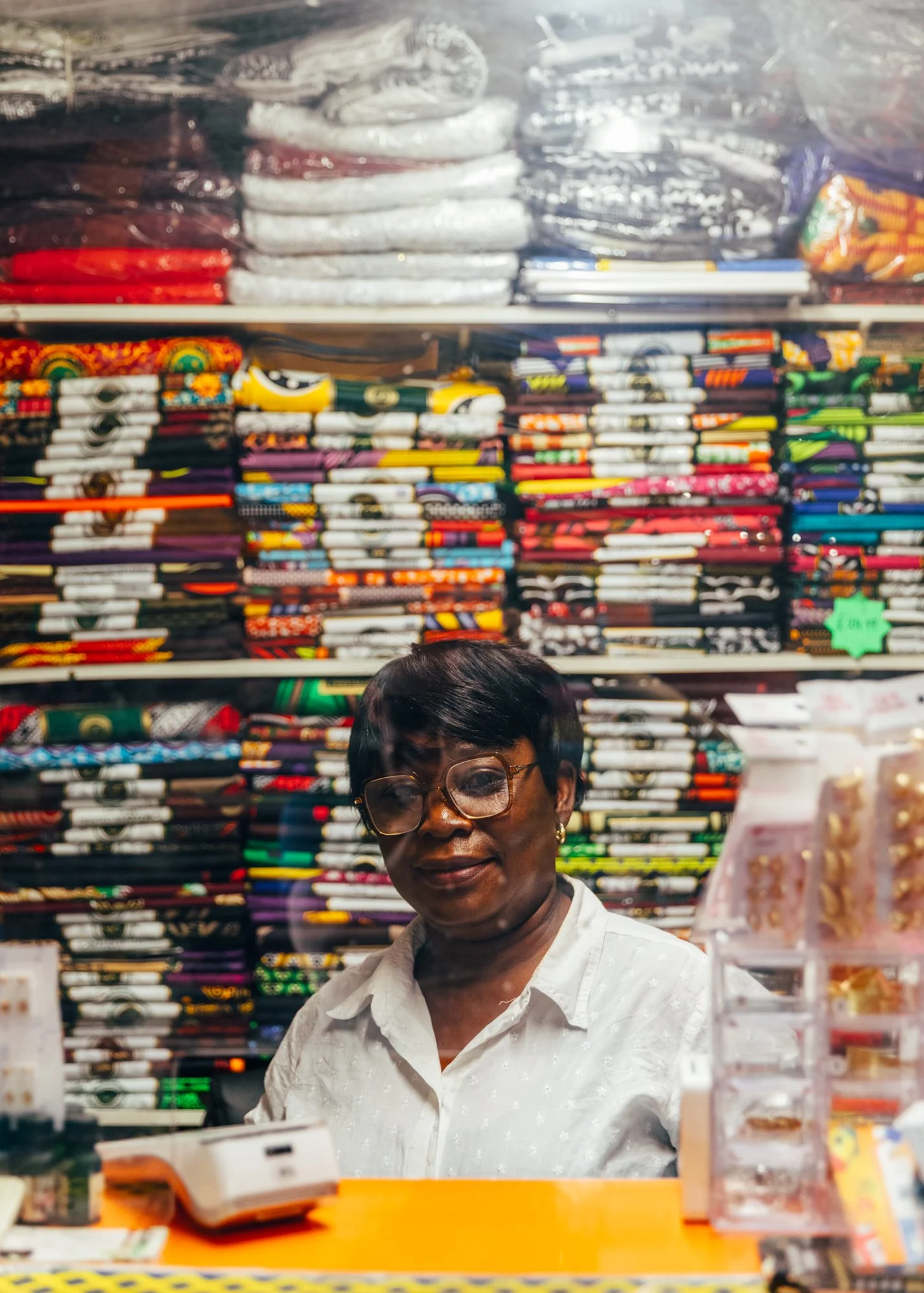 A woman wearing glasses stands behind a store counter with colorful fabric rolls and small jewelry items displayed around her, with more fabric in the background.