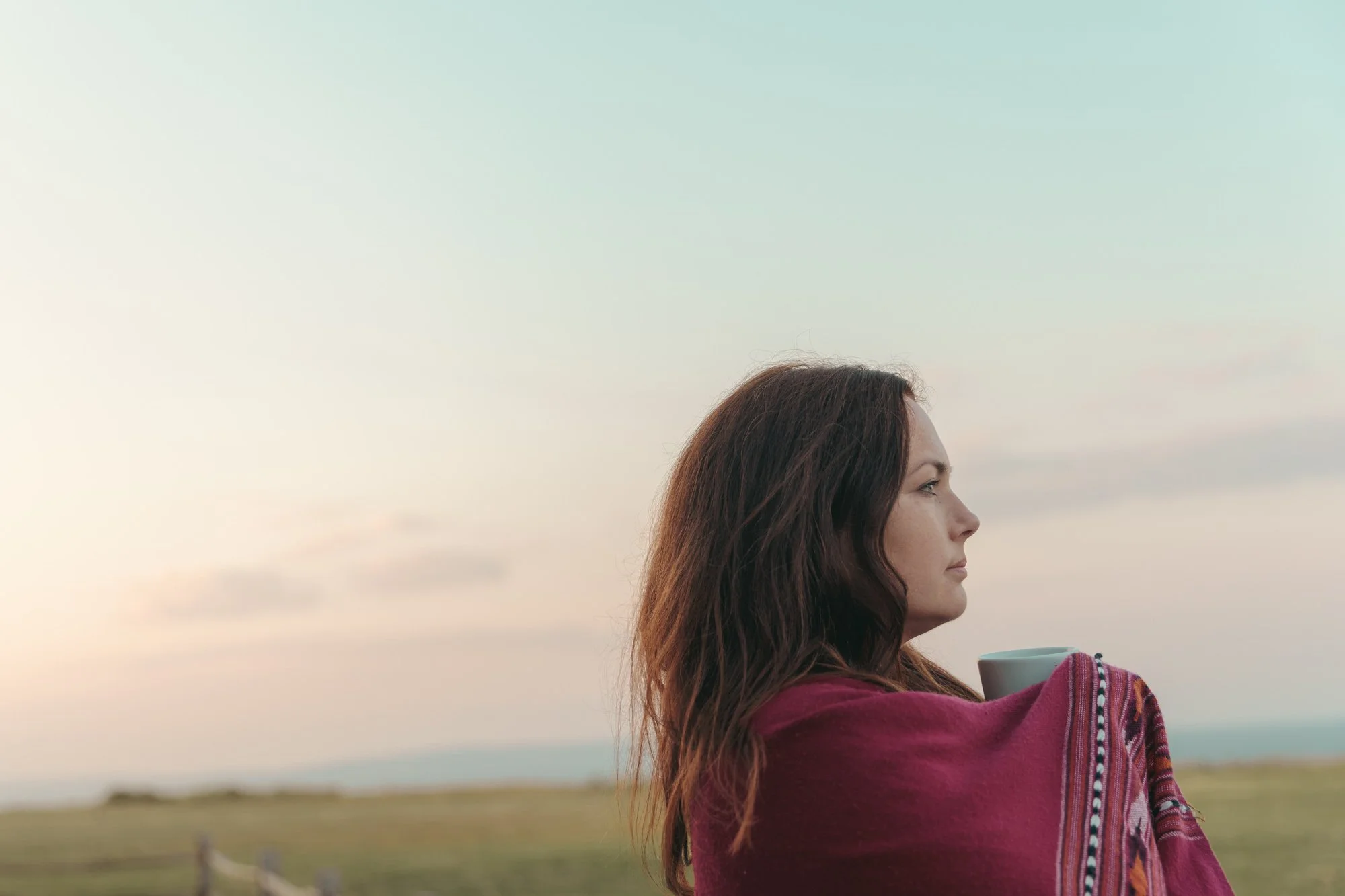 A woman with long brown hair in profile looking at a vast sky, wrapped in a colorful shawl, holding a mug, outdoors during dusk or dawn.