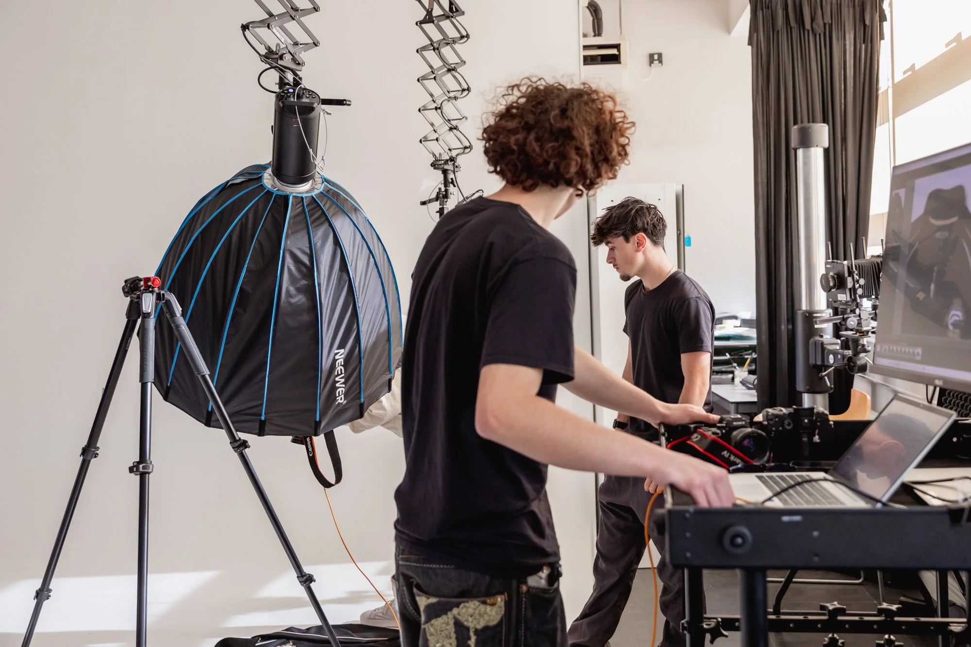 Two young men working with photography equipment in a studio, one adjusting a camera and the other looking at a computer screen, with studio lighting and backdrop in the background.
