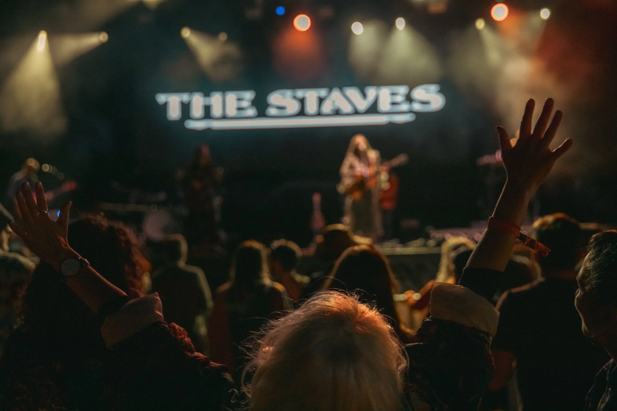 Audience at a concert raising their hands with a band performing on stage in the background with a large illuminated sign reading 'The Staves'.