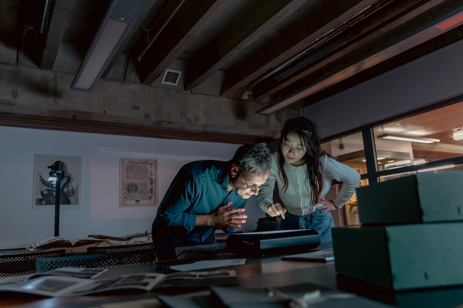 A woman and an older man look closely at a tablet on a desk in a dimly lit office or workspace, with papers and boxes on the table.