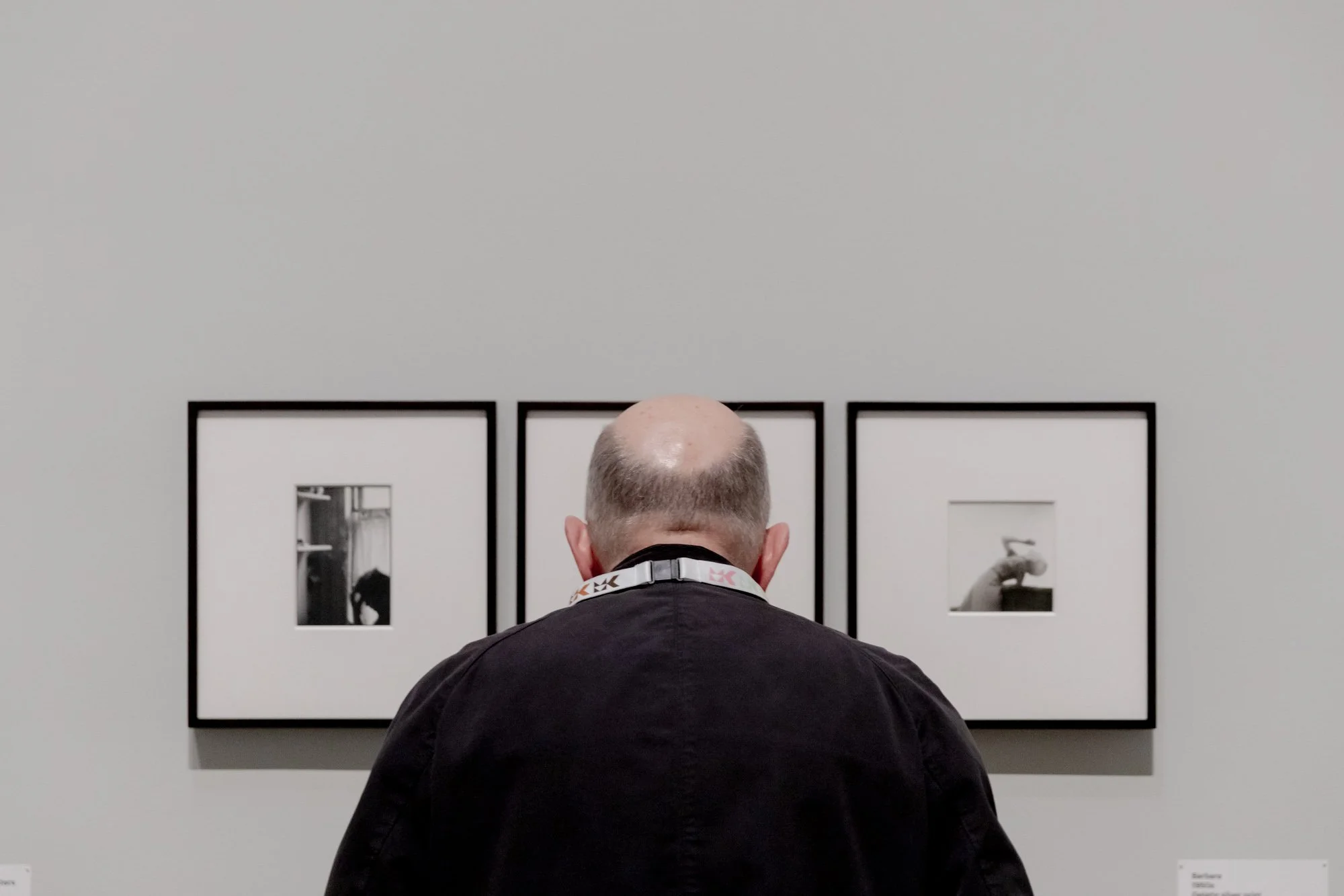 A man with a bald head wearing a black shirt looking at three black-and-white framed photographs on a white gallery wall.
