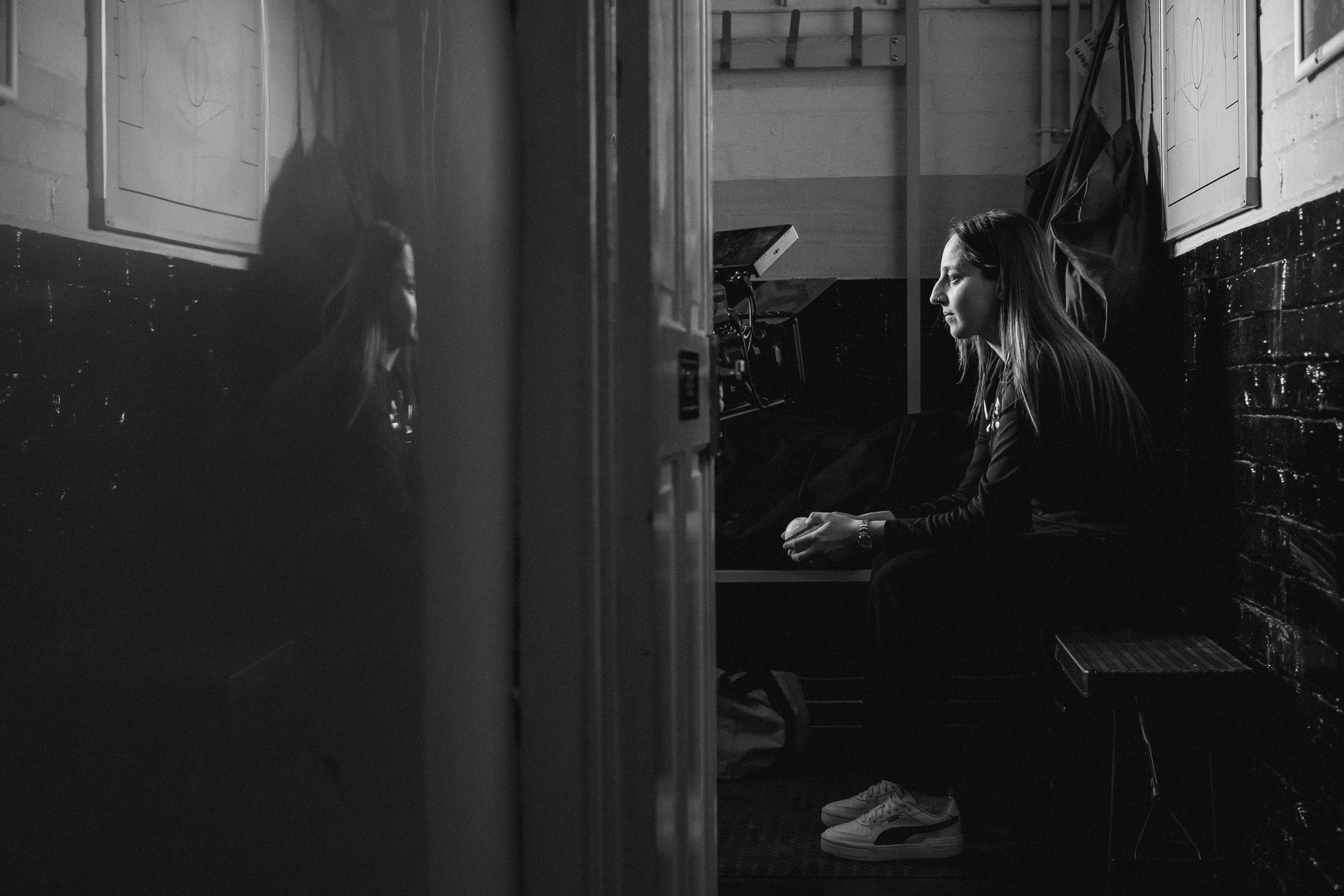 A woman sitting on a bench in a dimly lit room, with a serious expression, reflected in a mirror, black and white photo.