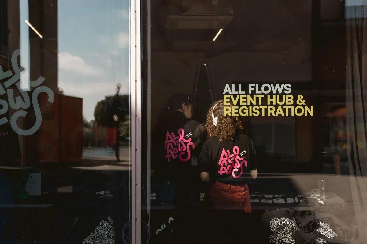 People attending an event, gathered behind a glass window with signage that reads "ALL FLOWS EVENT HUB & REGISTRATION," at what appears to be an outdoor conference or festival in the daytime.