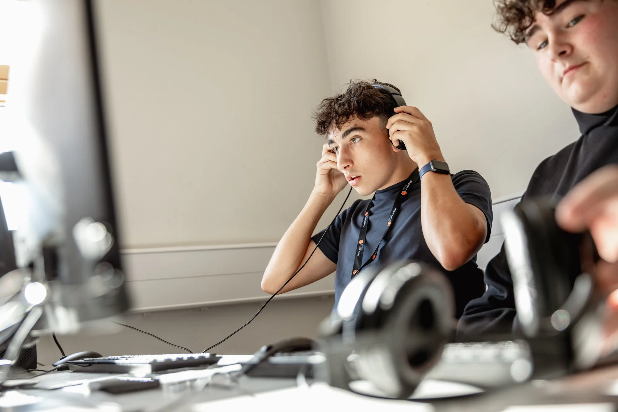 Young man wearing headphones at a desk, focused on a computer screen, with another person partially visible nearby.