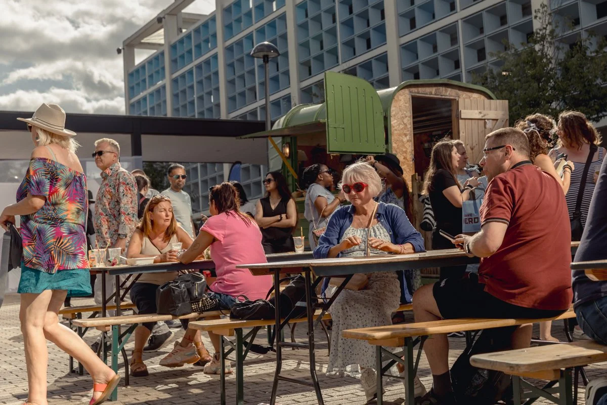 People sitting and standing at an outdoor event with food trucks and modern buildings in the background.