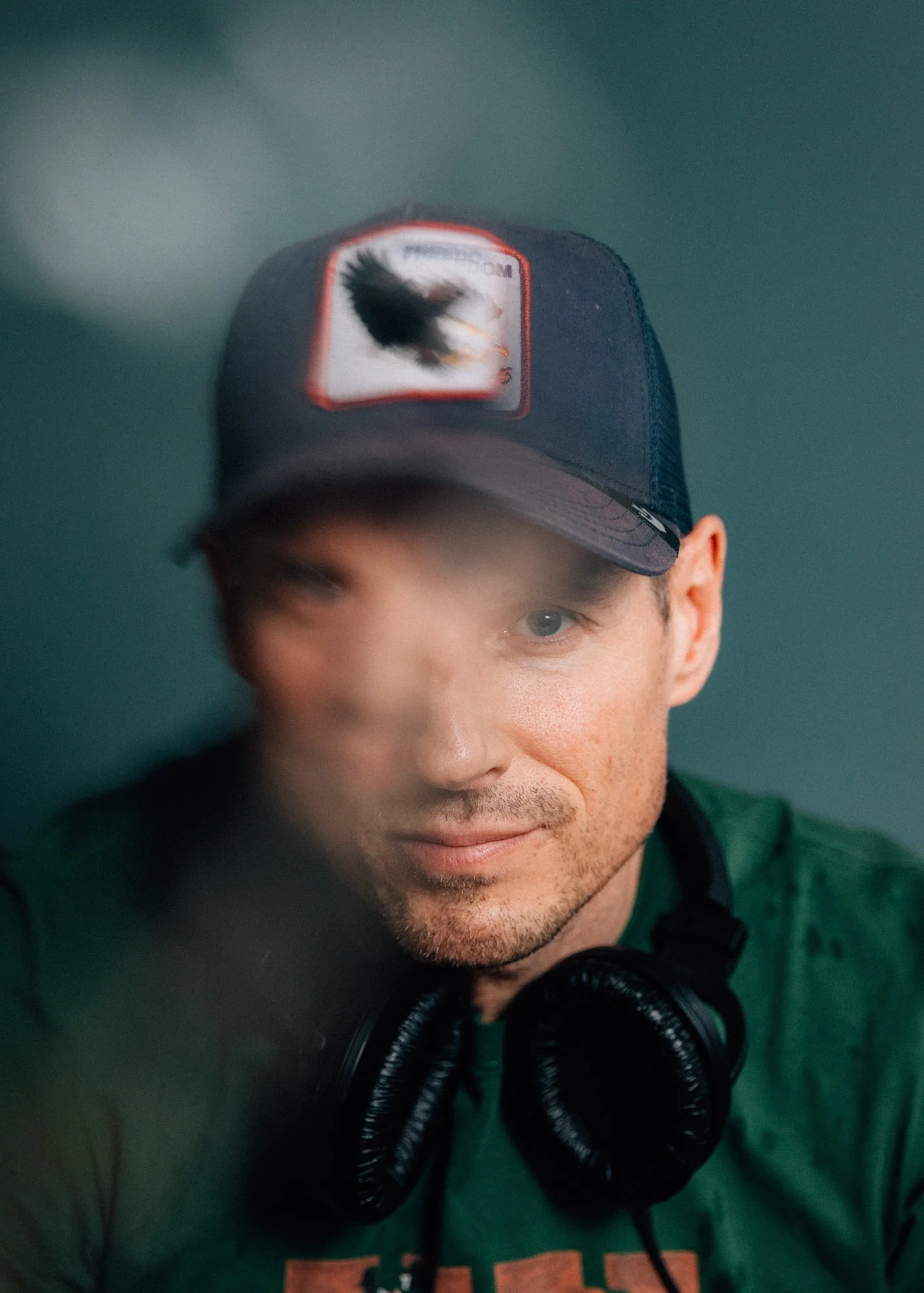 A man wearing a baseball cap, with headphones around his neck, looking into the camera through a glass surface, with a reflection slightly obscured in the foreground.