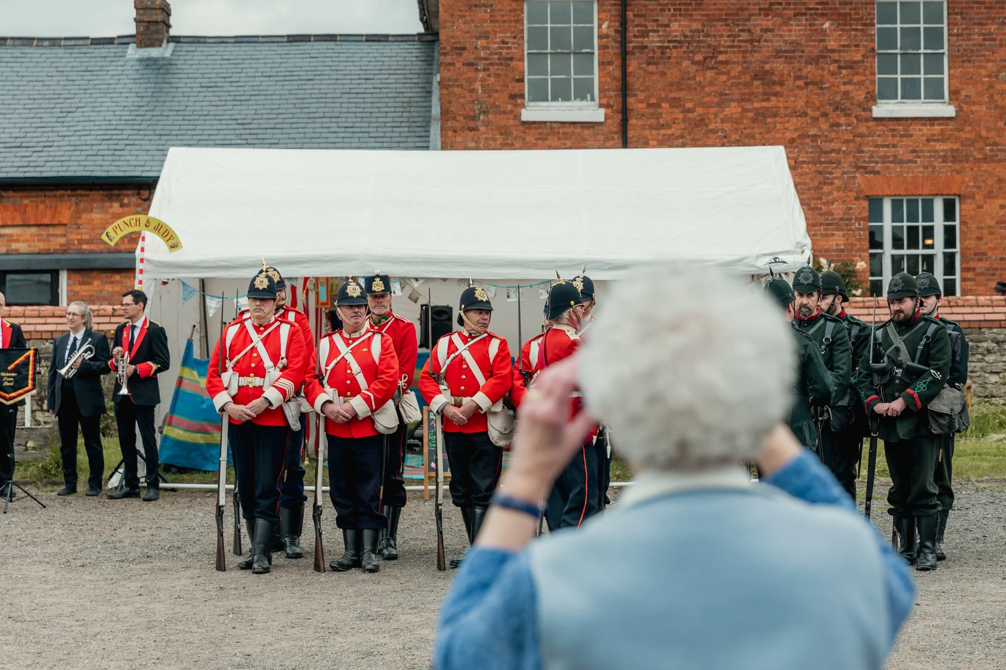 An elderly woman with white hair taking a photo of a group of men in historical military uniforms during a ceremony or reenactment outside a brick building.