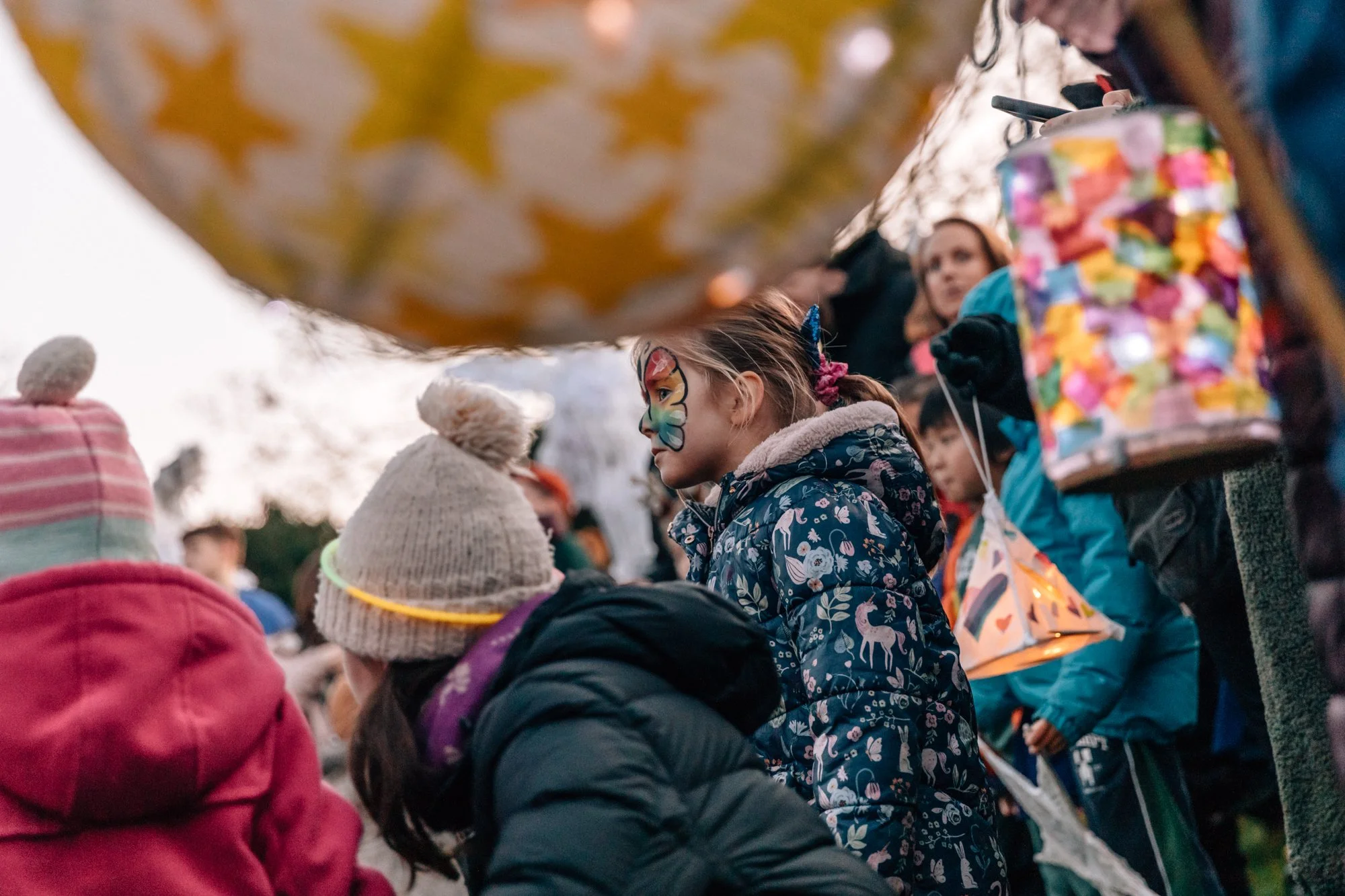 Group of children outdoors, including a girl with a butterfly face paint, surrounded by others in warm jackets and hats, some holding lanterns, during an evening event.