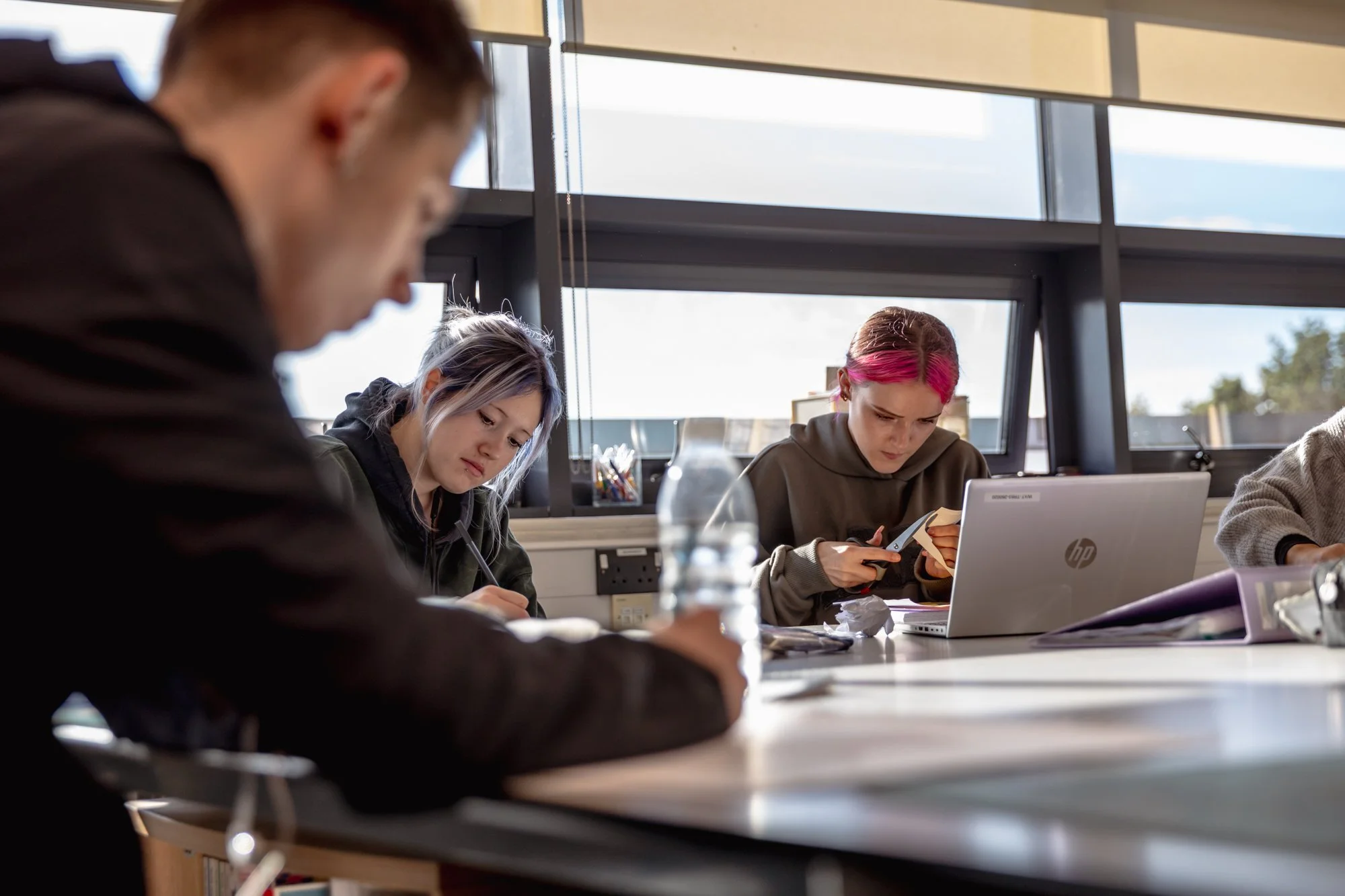 Students studying at a table in a classroom with large windows in the background.