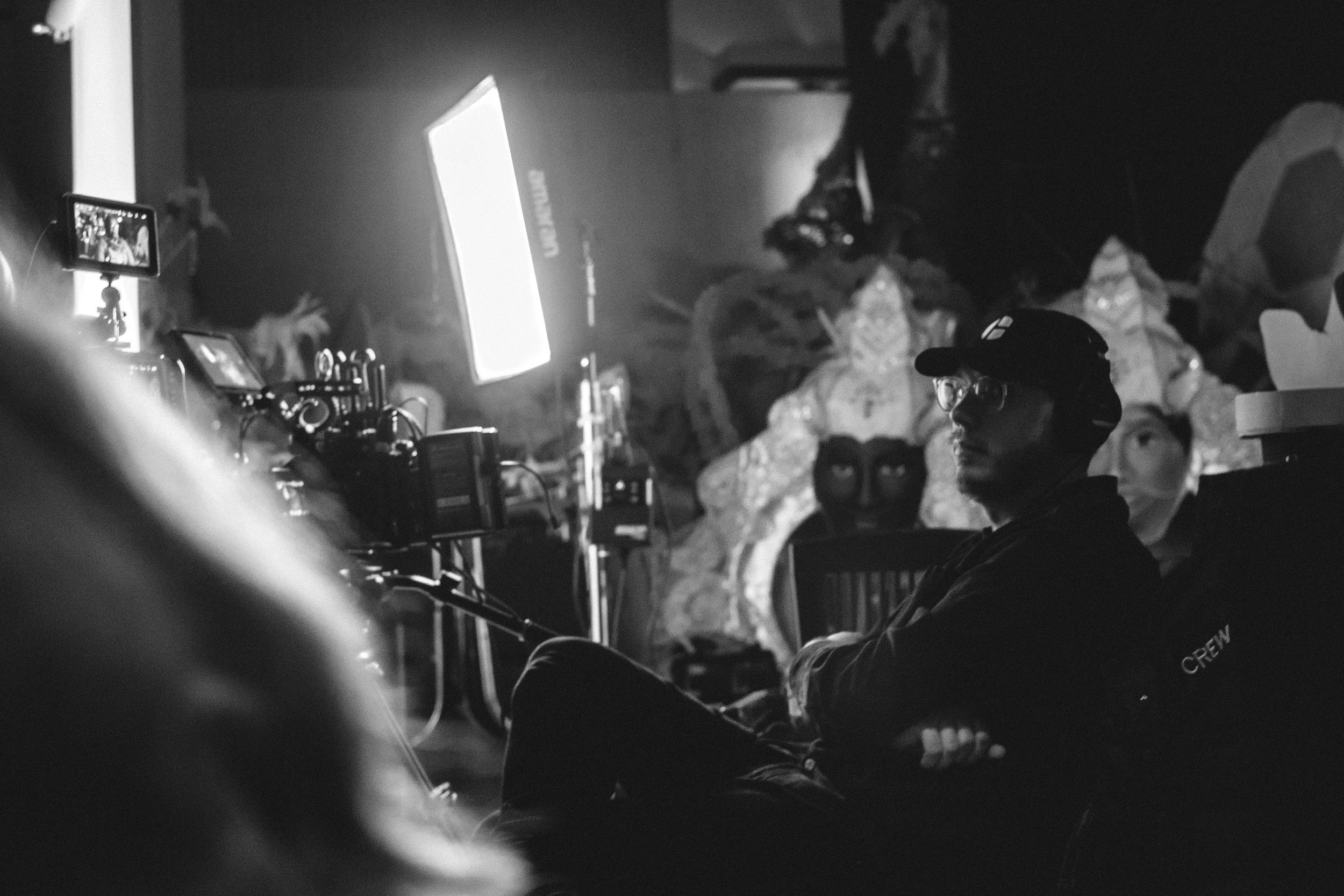 A person wearing glasses, a cap, and a black shirt with 'CREW' written on the sleeve, sitting in a dark room with professional lighting and camera equipment, behind a set with decorative masks and artwork.