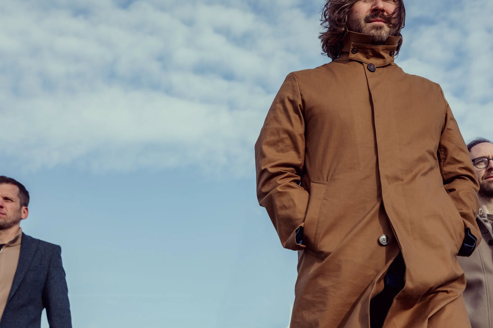 Three men standing outdoors against a cloudy sky, dressed in business casual attire. The man in the foreground wears a brown jacket, with his hands in his pockets, facing forward. The other two men are partially visible on each side.