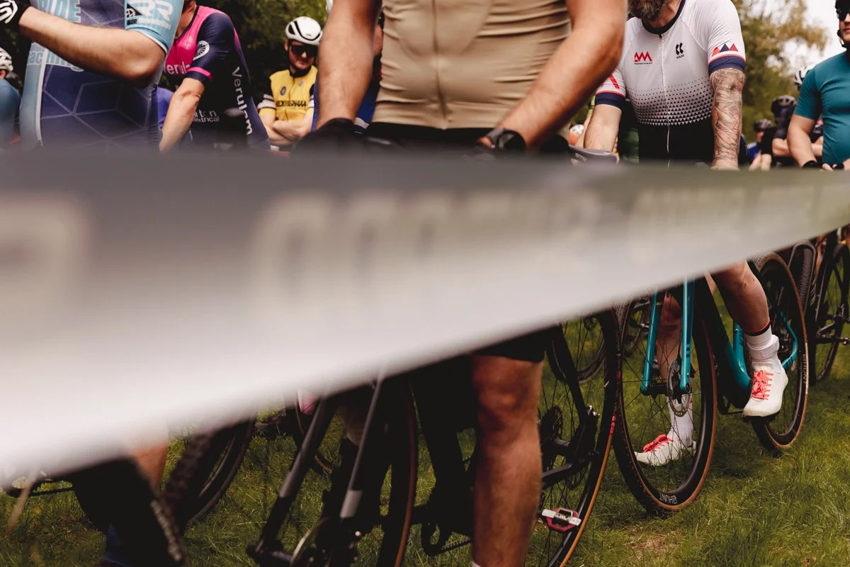 Cyclists preparing at the starting line, with a focus on their bikes and lower bodies, wearing cycling gear and shoes on a grassy outdoor area.
