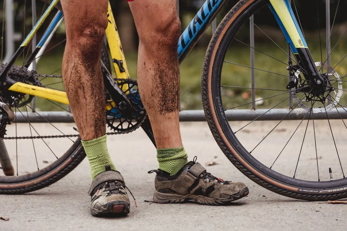 A cyclist's muddy legs and feet standing next to a yellow and blue mountain bike with disc brakes on a gravel path.