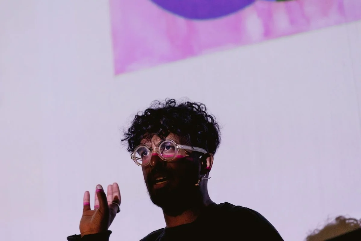 A man with curly hair and glasses speaking in front of a white wall with a partial pink and purple artwork above him.