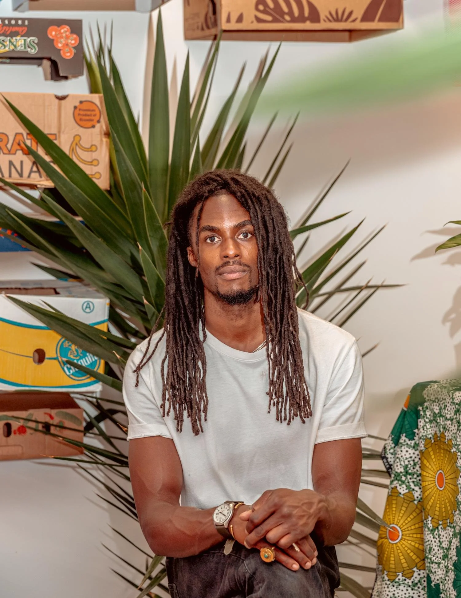 A man with long dreadlocks, wearing a white t-shirt and a wristwatch, sitting in front of large green plants and displayed shelves with boxes of snacks or food products.
