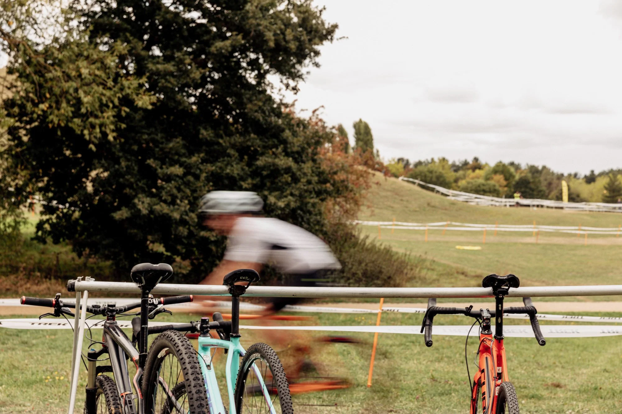 Blurred cyclist riding through a park with green grass, trees, and signs along the trail, as two bikes are parked in front of the fence.