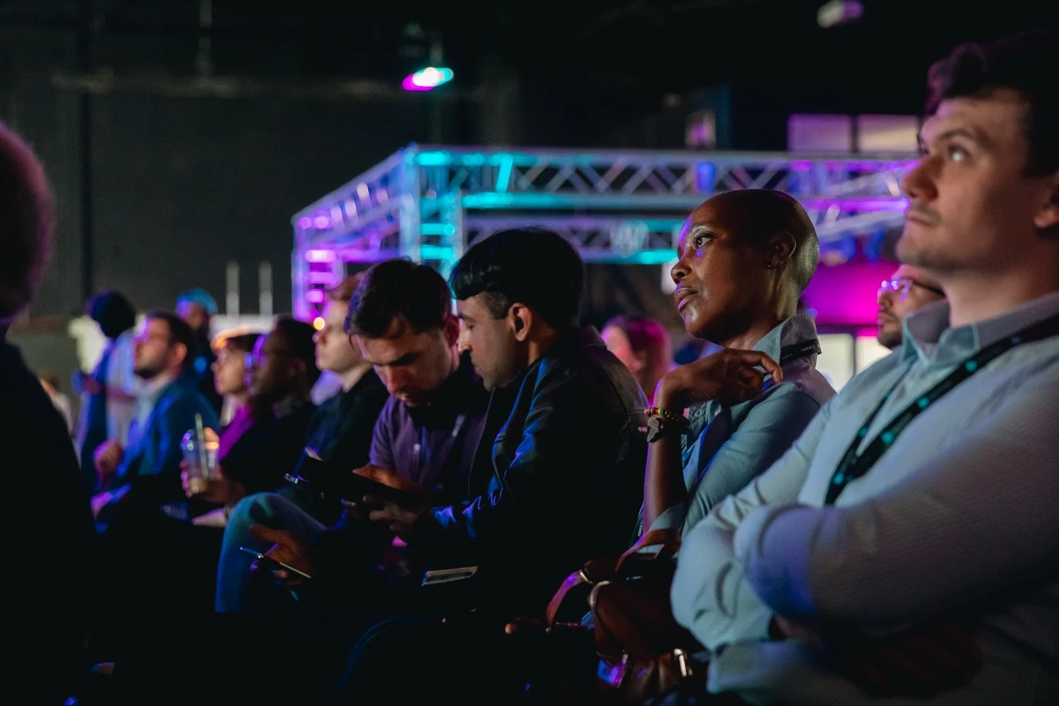 Audience members attentively listening at an indoor event with colorful stage lighting.