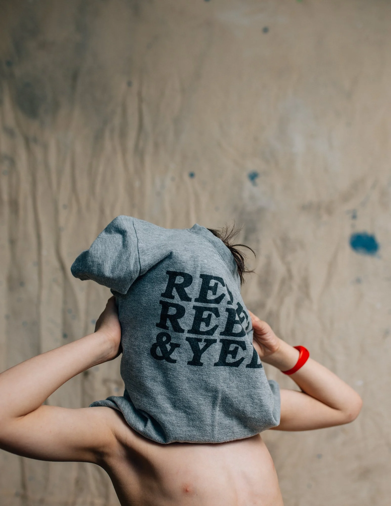 A shirtless person holding a gray hoodie with text on the back, covering their face, with a wooden wall background.