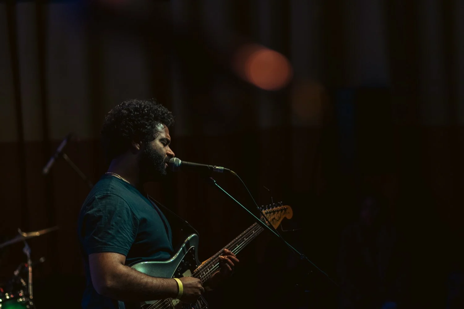 A man with curly hair and a beard playing an electric guitar and singing into a microphone on stage, dimly lit with some blurred lights in the background.