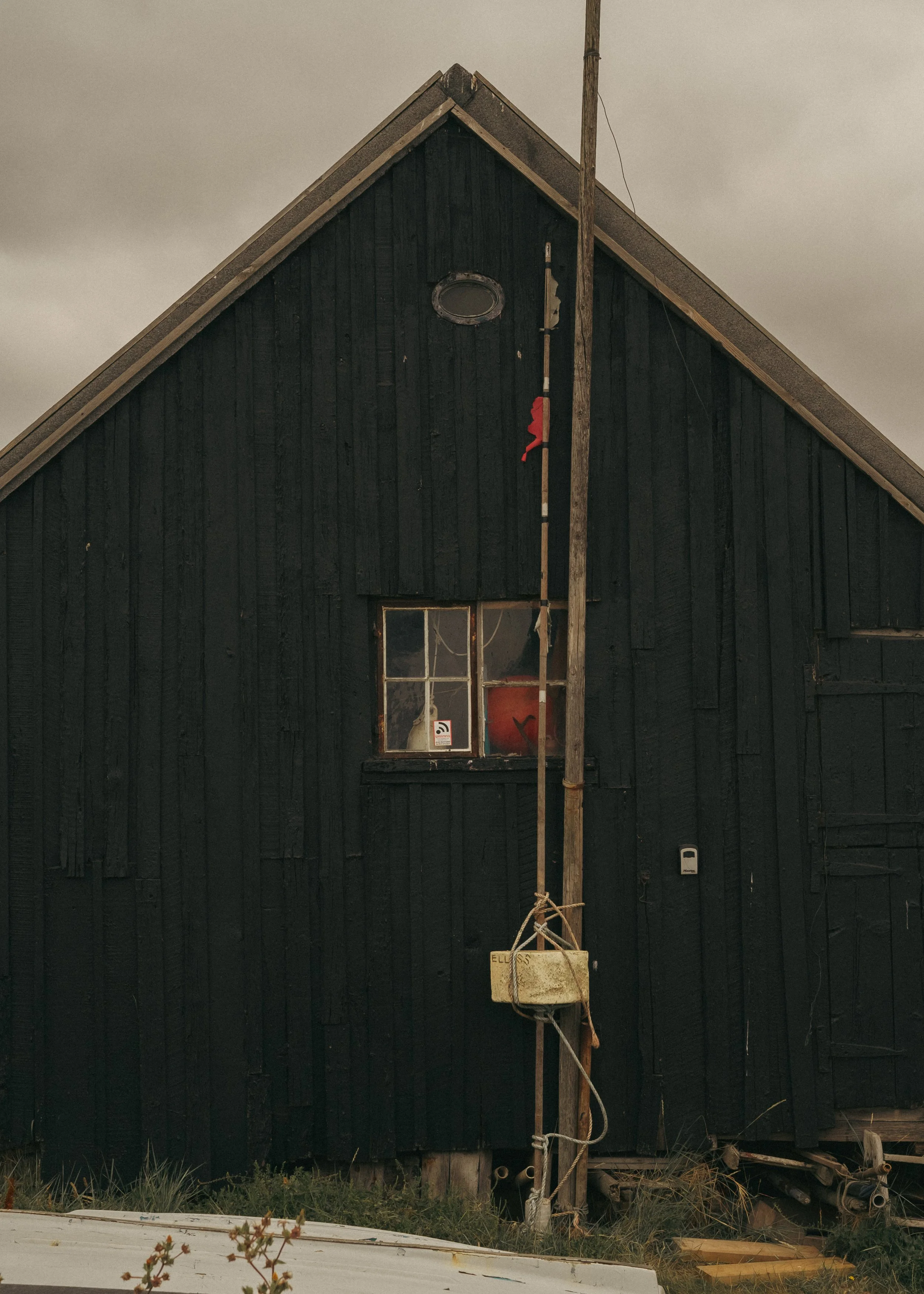 A black wooden house with a small window, a sloped roof, and a wooden pole in front, tied with ropes, under a cloudy sky.