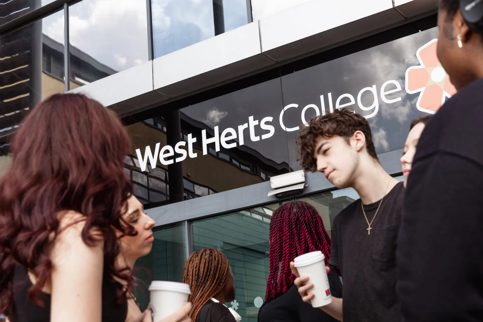 A group of diverse students talking and holding coffee cups outside West Herts College building.