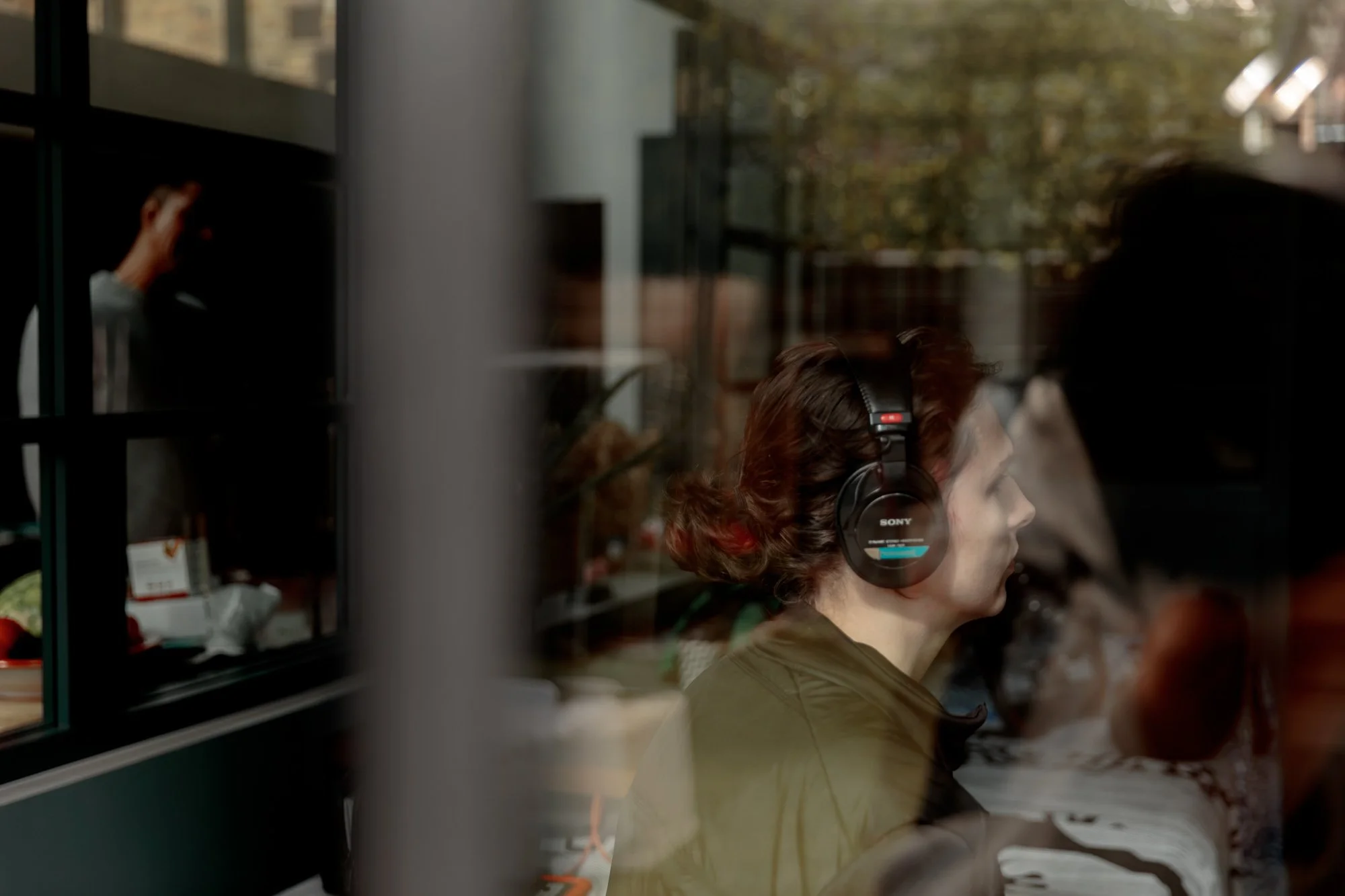 A woman with brown hair and headphones sitting inside, seen through a glass window, with a reflection of her profile and an outdoor scene with trees in the background.
