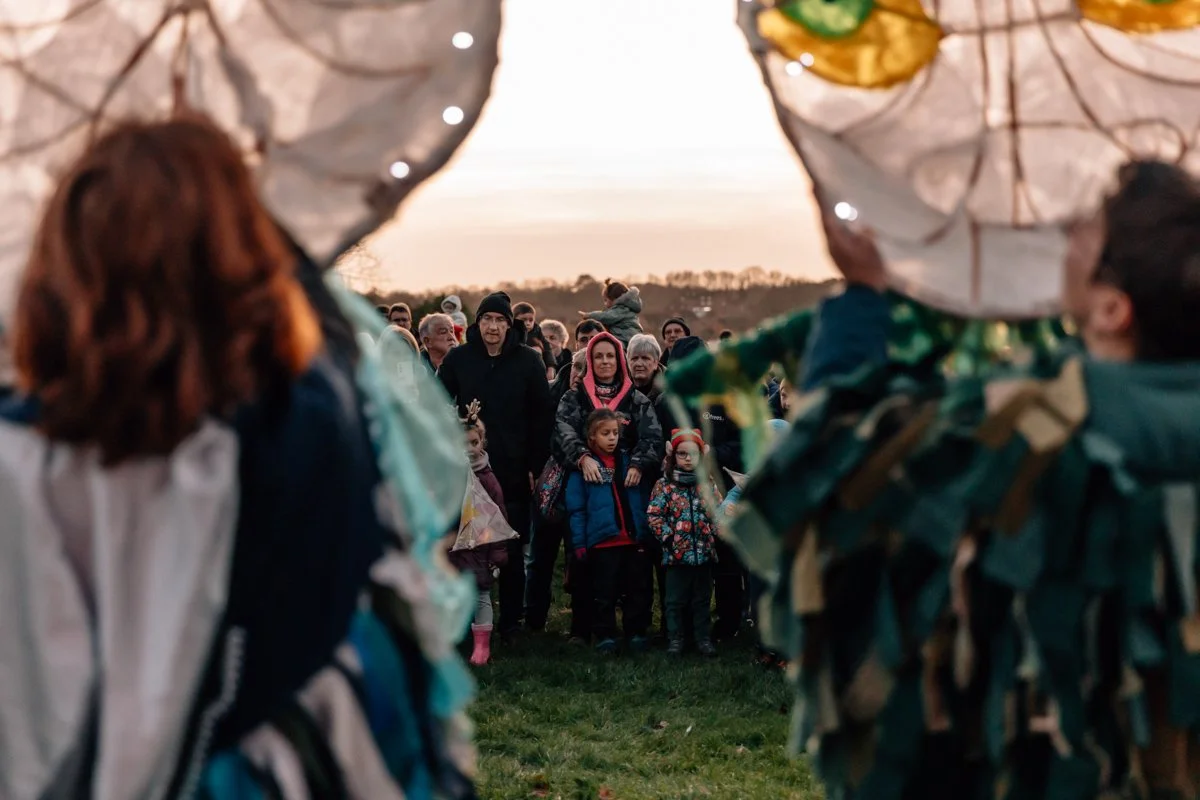 A crowd of people, including children, gathered outdoors at sunset, seen through the heads of two people carrying large fabric balloons.