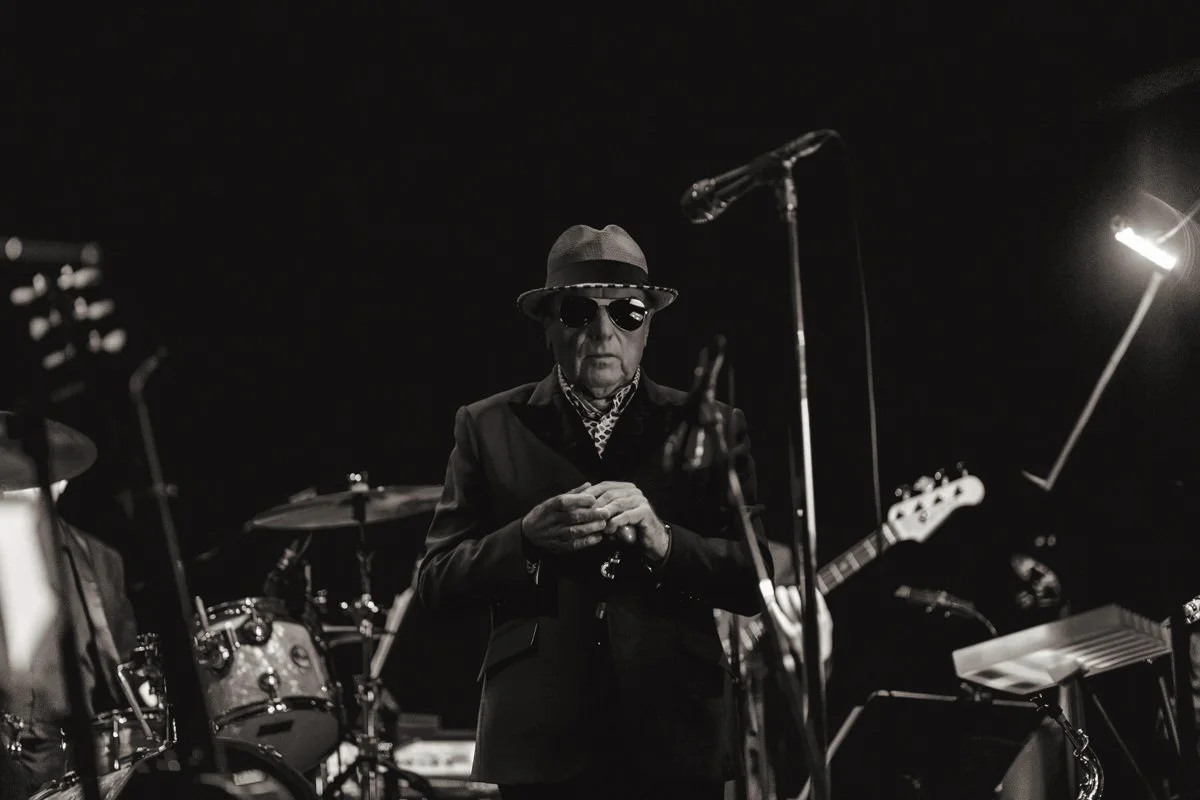 A black and white photo of a man in dark sunglasses and a hat, standing on a stage with musical instruments, including drums and a guitar, holding an object in his hands.