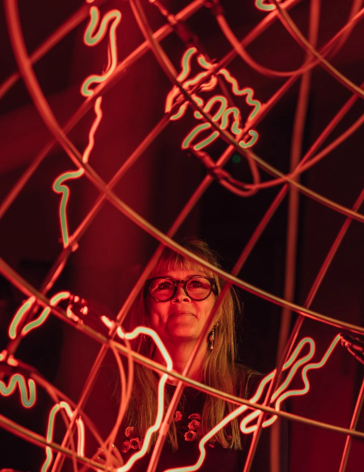A woman with glasses and long hair standing behind a large illuminated globe-shaped neon sign with red and green lights.