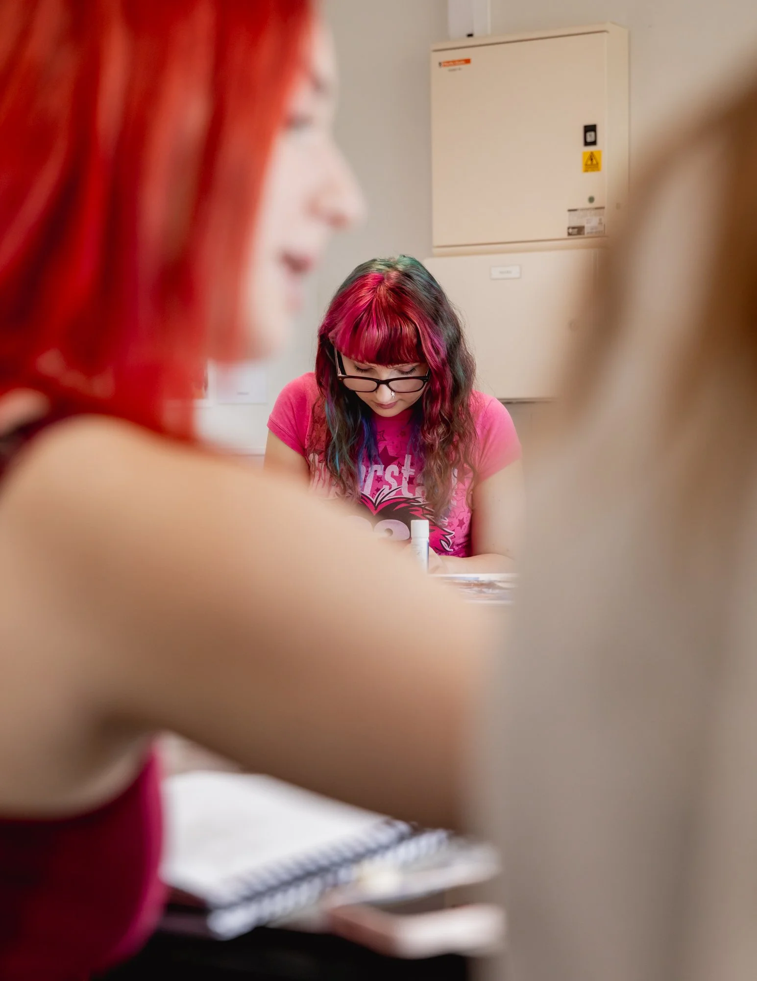A woman with dark pink and teal hair wearing glasses and a pink T-shirt sitting at a table, seen through blurred foreground people with similar hair colors.