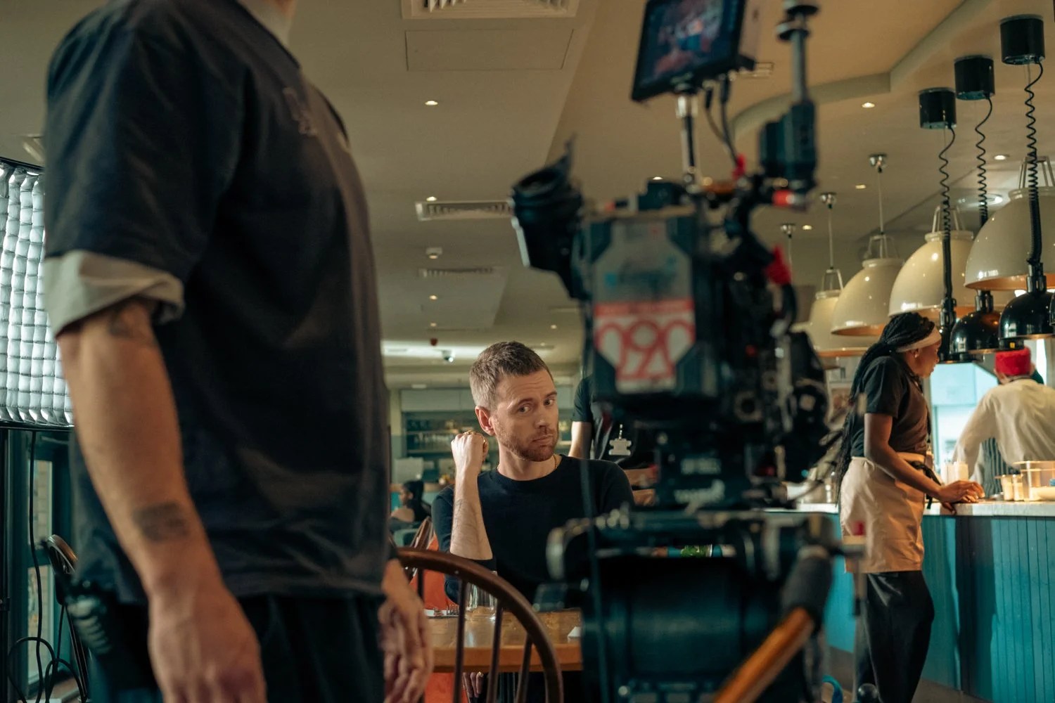 A film crew is filming in a restaurant. A man with a beard and black shirt is sitting at a table, looking at the camera. A crew member is standing nearby, partially visible. Two women are working behind the counter, one wearing a black shirt and apro