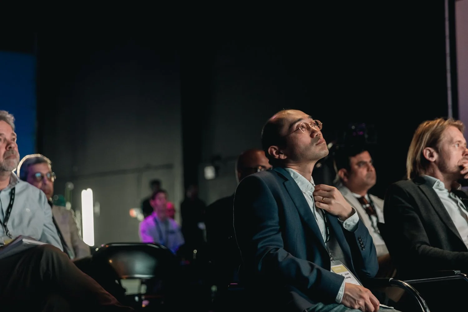 Attendees at a conference or seminar listening attentively, seated in rows, some wearing identification lanyards, in a darkly lit room with stage lighting.