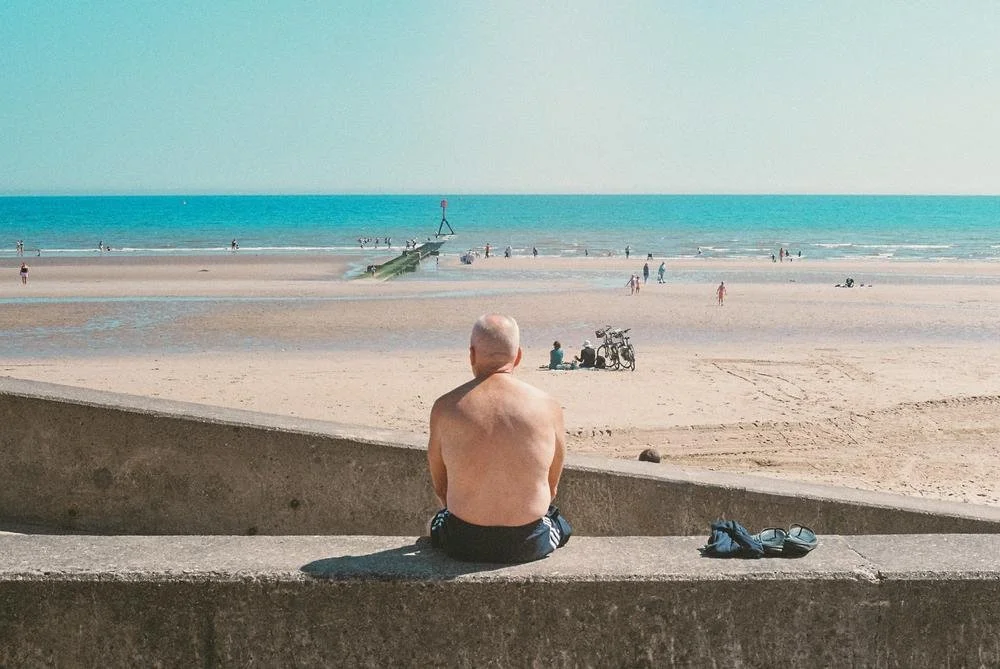 A man sitting on a concrete ledge at the beach, facing the ocean with a pier extending into the water, surrounded by beachgoers and bicycles.