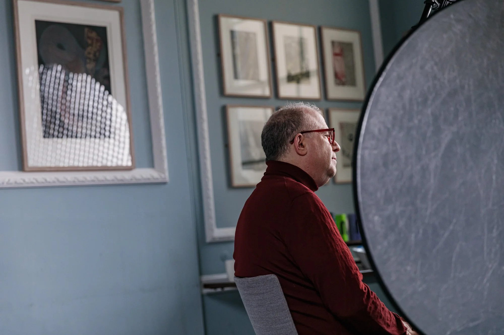 A man with glasses and a red turtleneck sitzt vor einem grauen Hintergrund in einem Raum mit mehreren Bildern an der Wand.