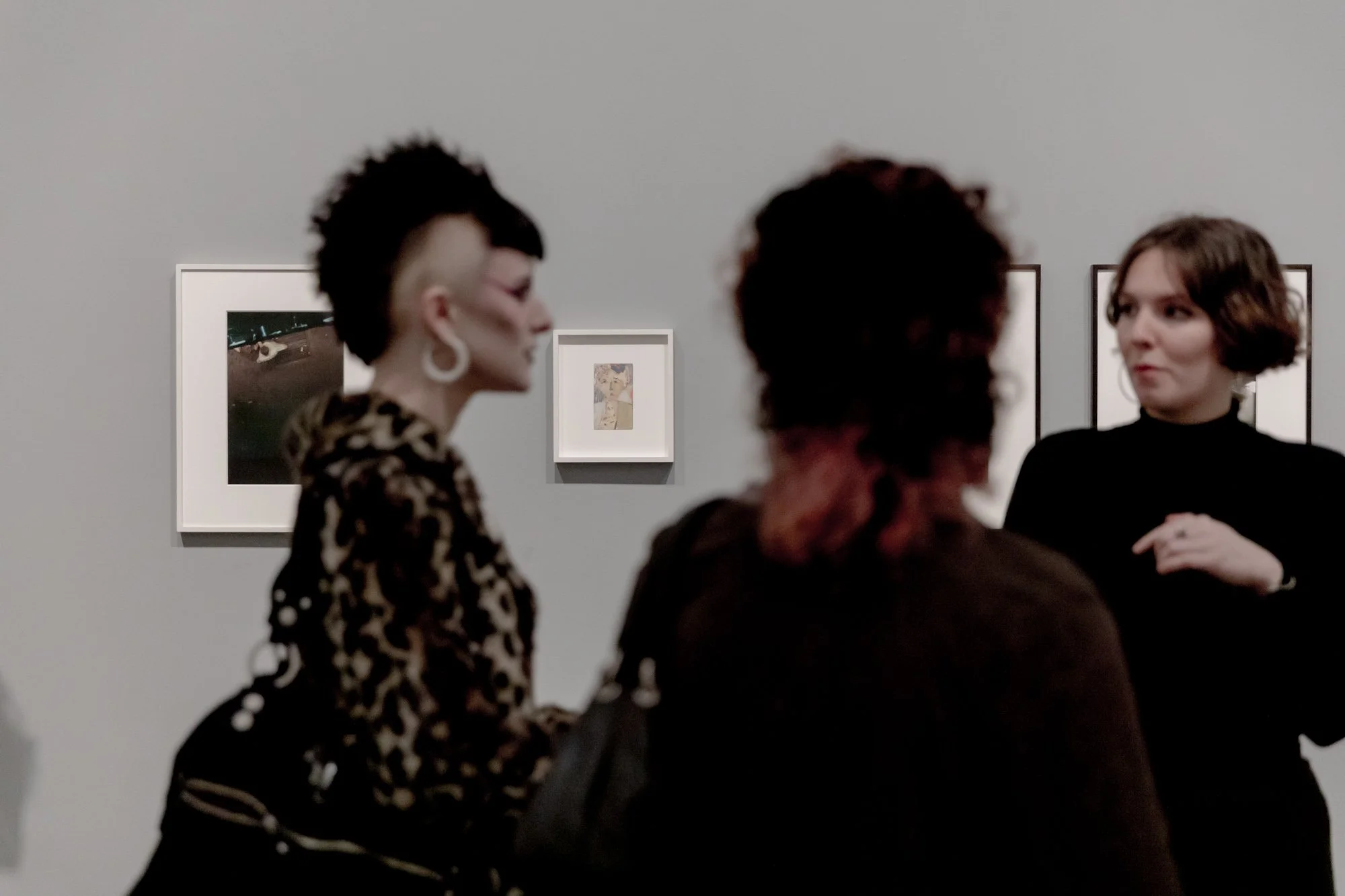 Three women engaged in conversation at an art gallery, with framed artworks hanging on a gray wall behind them.