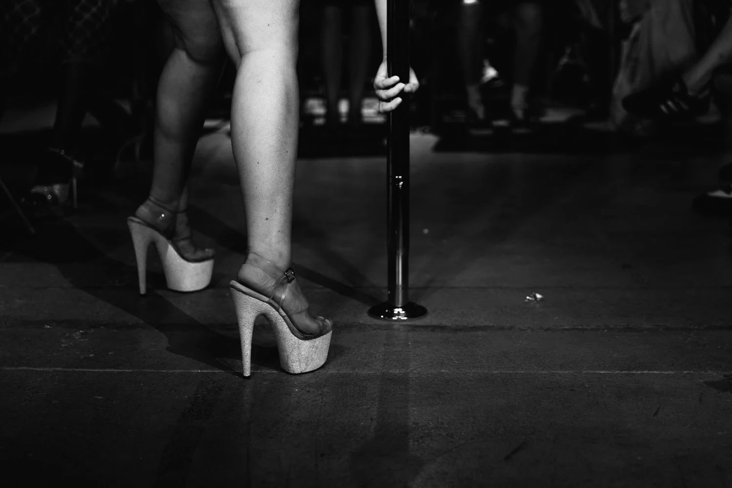 A person standing on high heels, holding onto a pole, with an audience in the background, in a black and white photo.