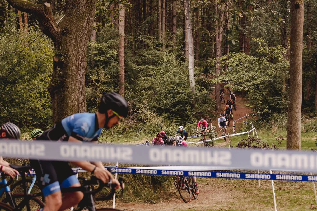 Cyclists riding mountain bikes through a forested trail during a race, with some riders pushing and others riding uphill, surrounded by trees and greenery.