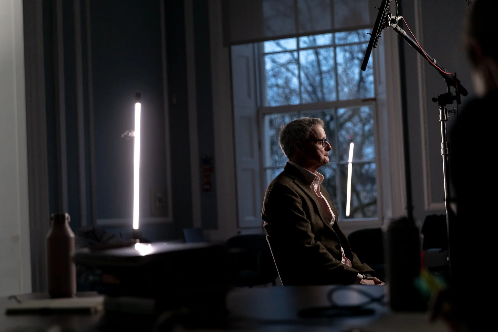 A man with glasses sitting in profile during an interview or video shoot, indoors with large windows showing an outdoor scene with leafless trees, illuminated by vertical studio lights.