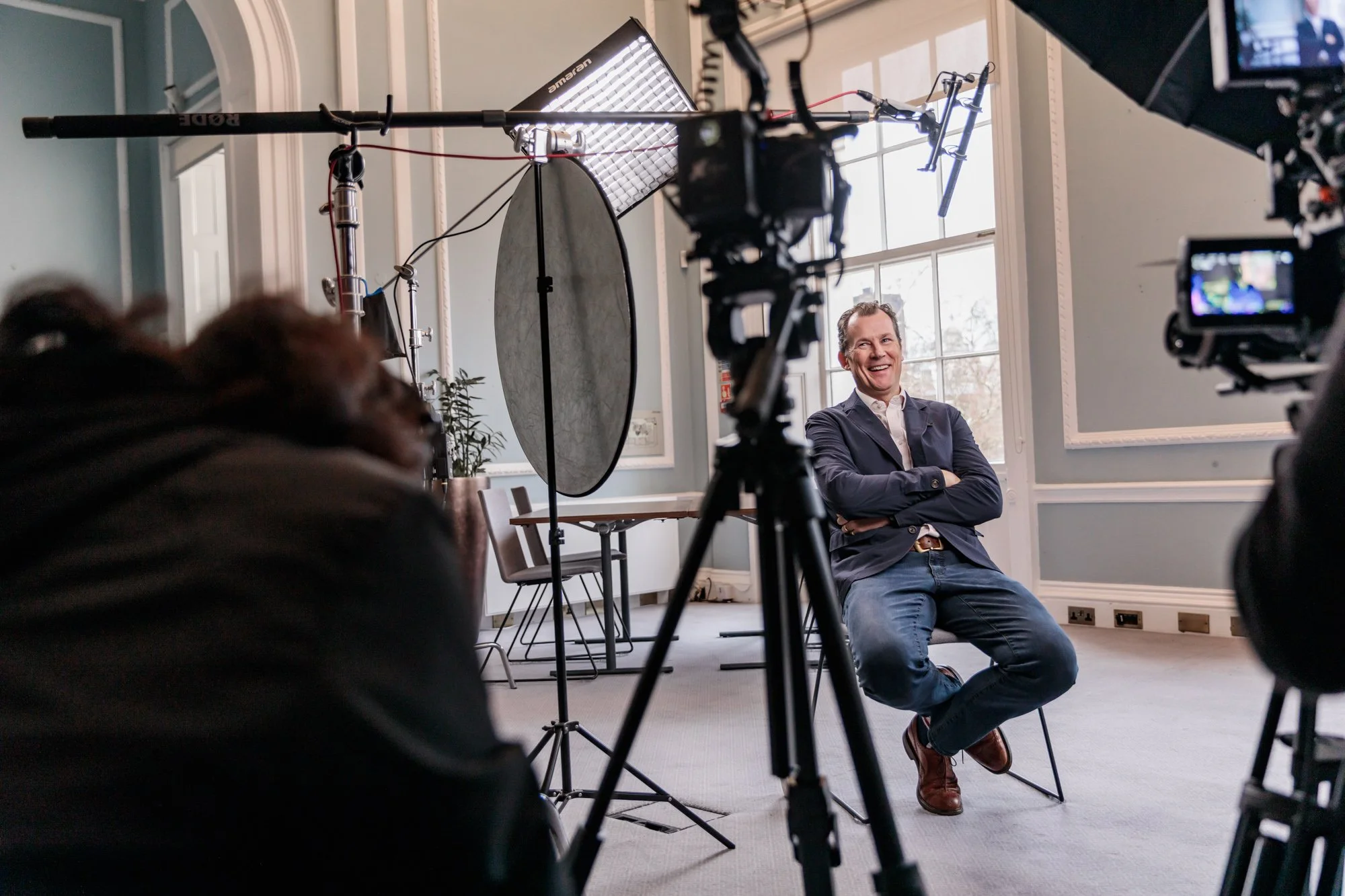 A man sitting on a chair during an interview or filming session in a well-lit room with large windows, surrounded by camera equipment and lighting tools.