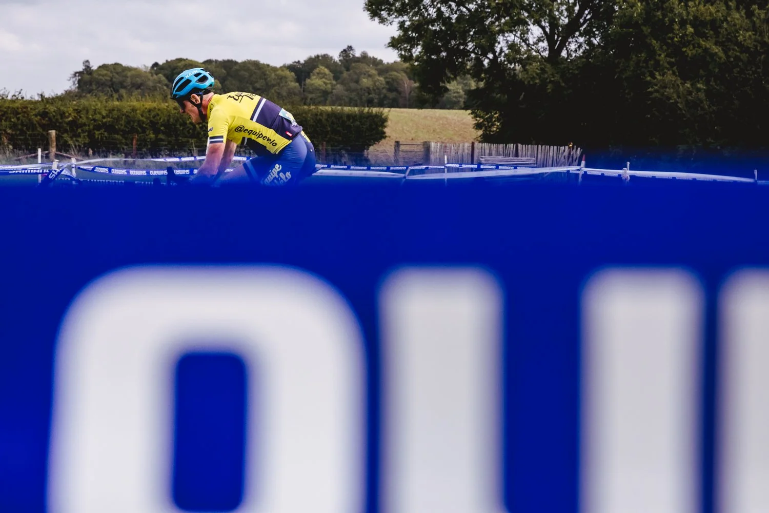 A cyclist in a yellow jersey and blue helmet riding on a mountain bike in a rural outdoor setting, with a fence, trees, and cloudy sky in the background.