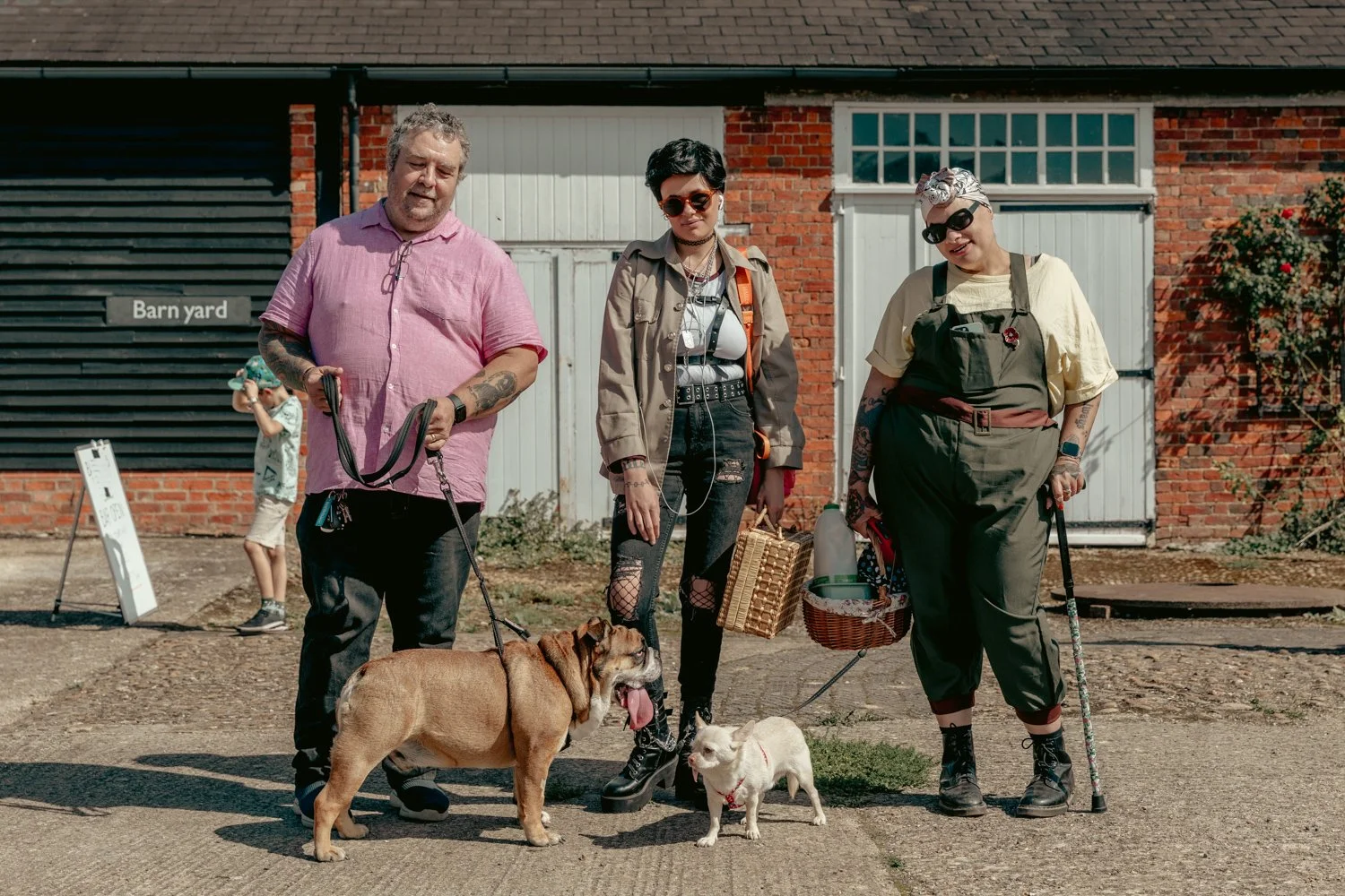 Three people standing in front of a brick building with white garages, holding dogs and leashes, with a boy in the background.
