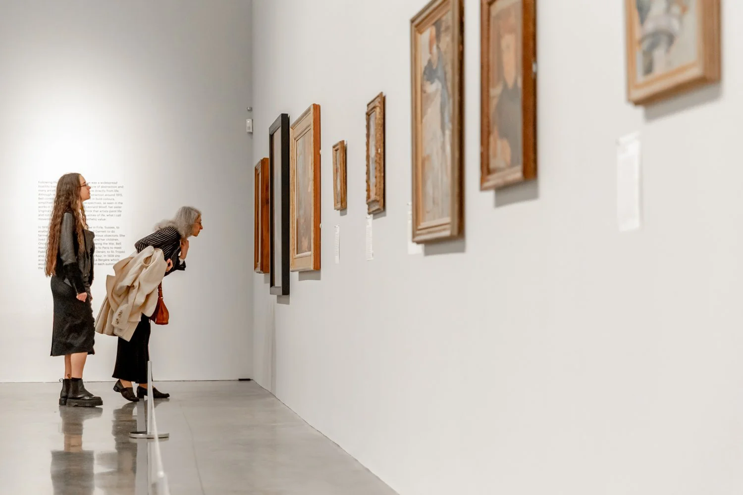 Two women look at artwork displayed on a white gallery wall, with informational placards beneath each piece