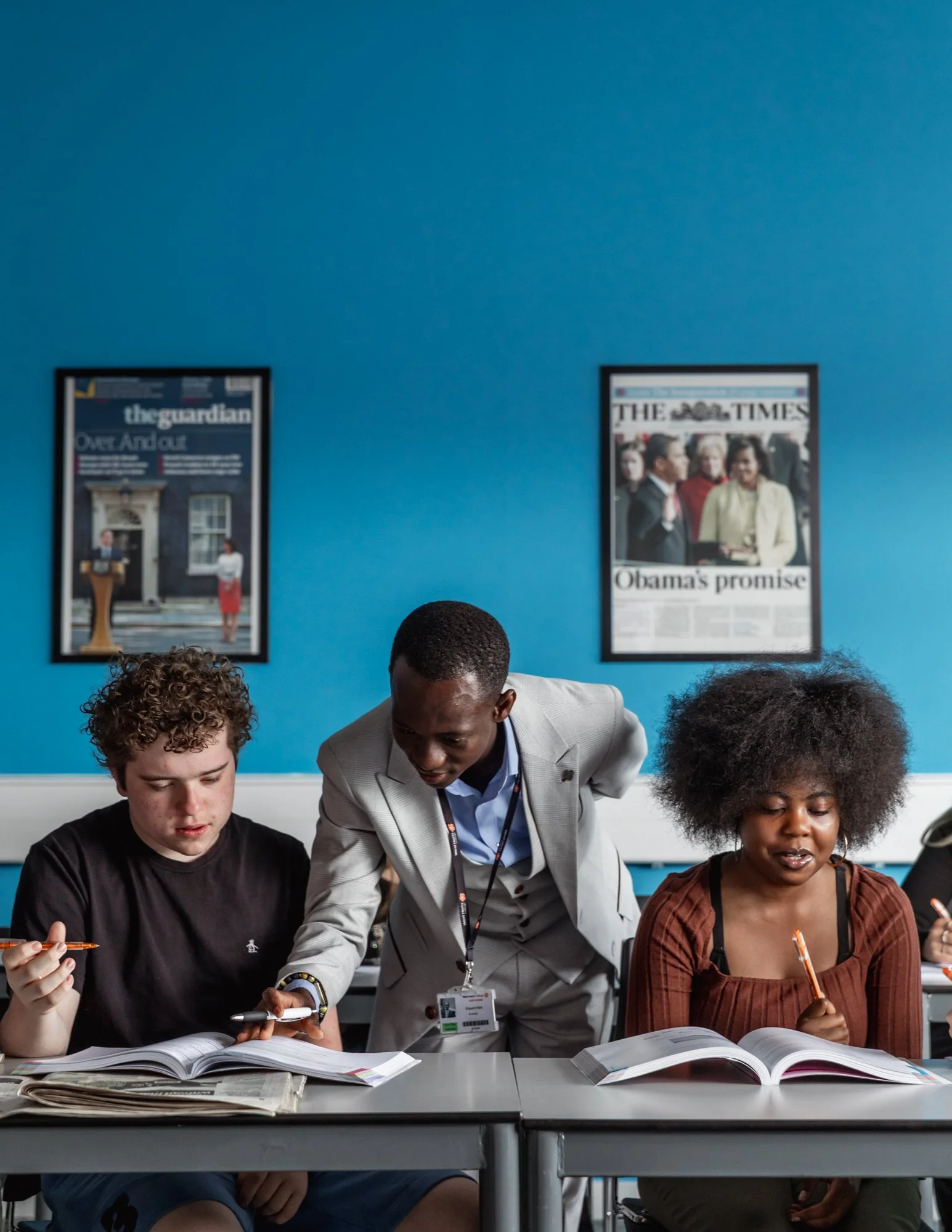 A teacher assisting two students with their studies in a classroom with blue walls and newspapers on the wall.