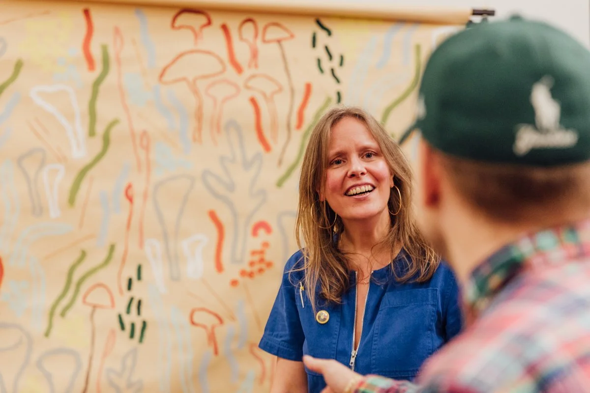 Two women talking in front of an abstract mural with various colored lines and shapes.