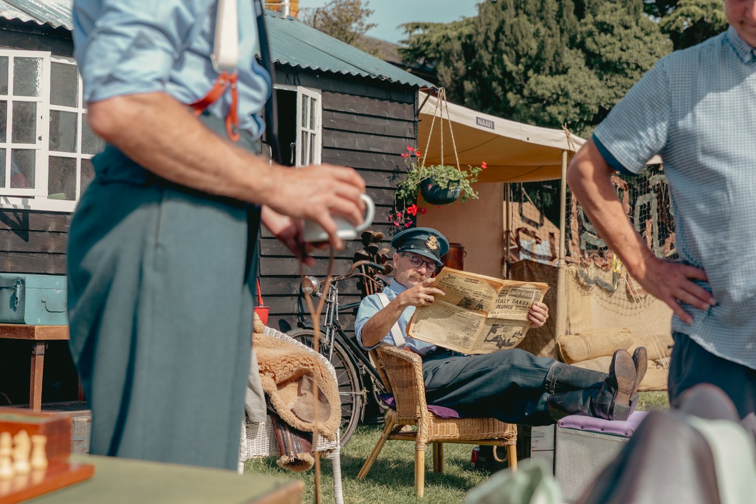 A man dressed as a police officer sitting in a wicker chair and reading a newspaper, while two other men stand nearby in a garden setting with a shed, bicycle, and hanging flowers in the background.
