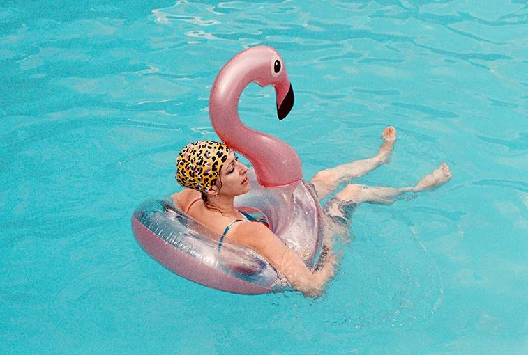 Woman in a swimming pool wearing a leopard print swim cap and orange swimsuit, sitting in an inflatable pink flamingo pool float.