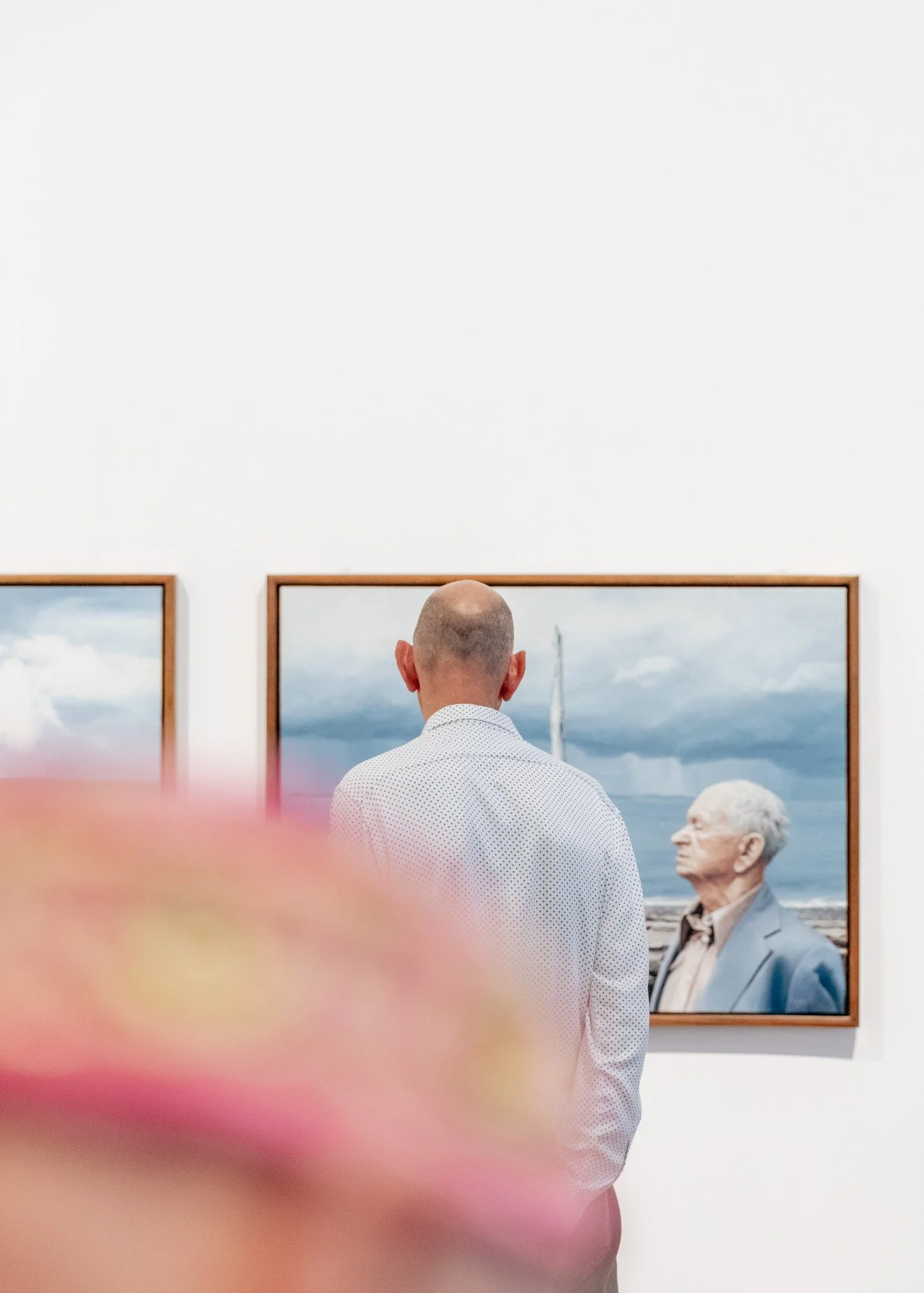 A person with a shaved head and white polka-dot shirt viewing paintings in an art gallery, with a blurred pink object in the foreground.