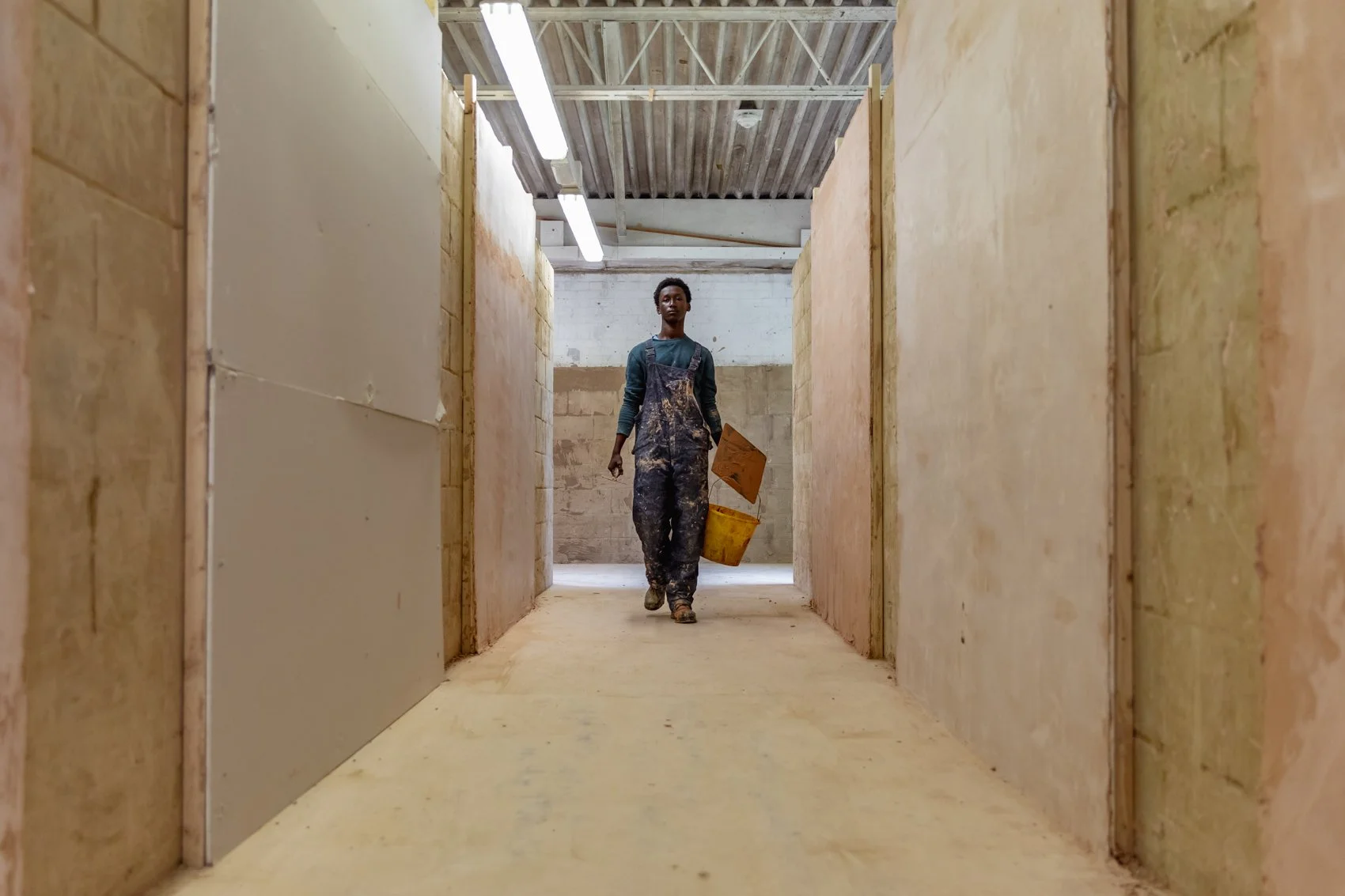 A person walking through a construction site corridor with insulation and drywall being installed, carrying a bucket and a board.